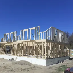 Surowy szkielet dwukondygnacyjnego domu z drewna, widoczne elementy konstrukcyjne, podmurówka z białych płyt izolacyjnych, błękitne niebo w tle.