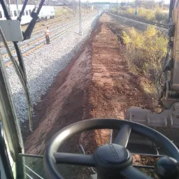 Widok z kabiny koparki na przygotowany nasyp ziemny wzdłuż torów kolejowych, z pracownikiem w odblaskowej kamizelce stojącym obok pociągu w tle.