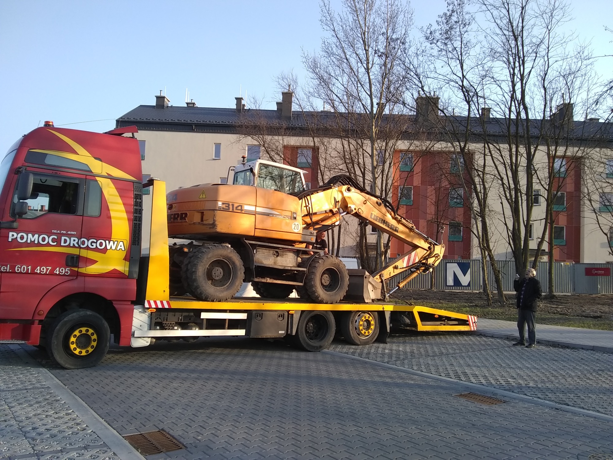 Żółta koparka LIEBHERR 314 na lawecie czerwonego samochodu pomocy drogowej z numerem telefonu 601 497 495, w tle budynek w trakcie budowy i mężczyzna w ciemnej kurtce.