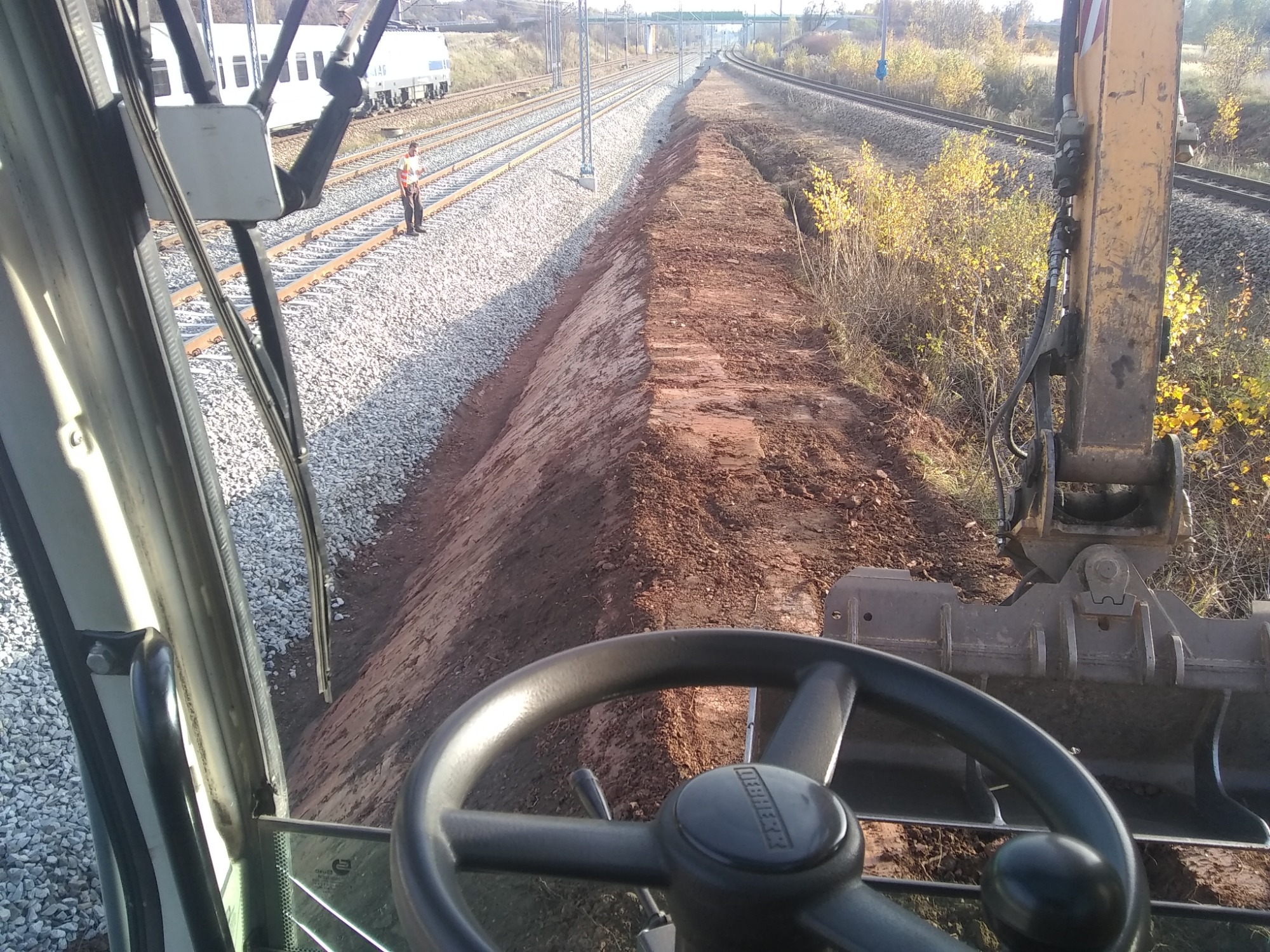 Widok z kabiny koparki na przygotowany nasyp ziemny wzdłuż torów kolejowych, z pracownikiem w odblaskowej kamizelce stojącym obok pociągu w tle.