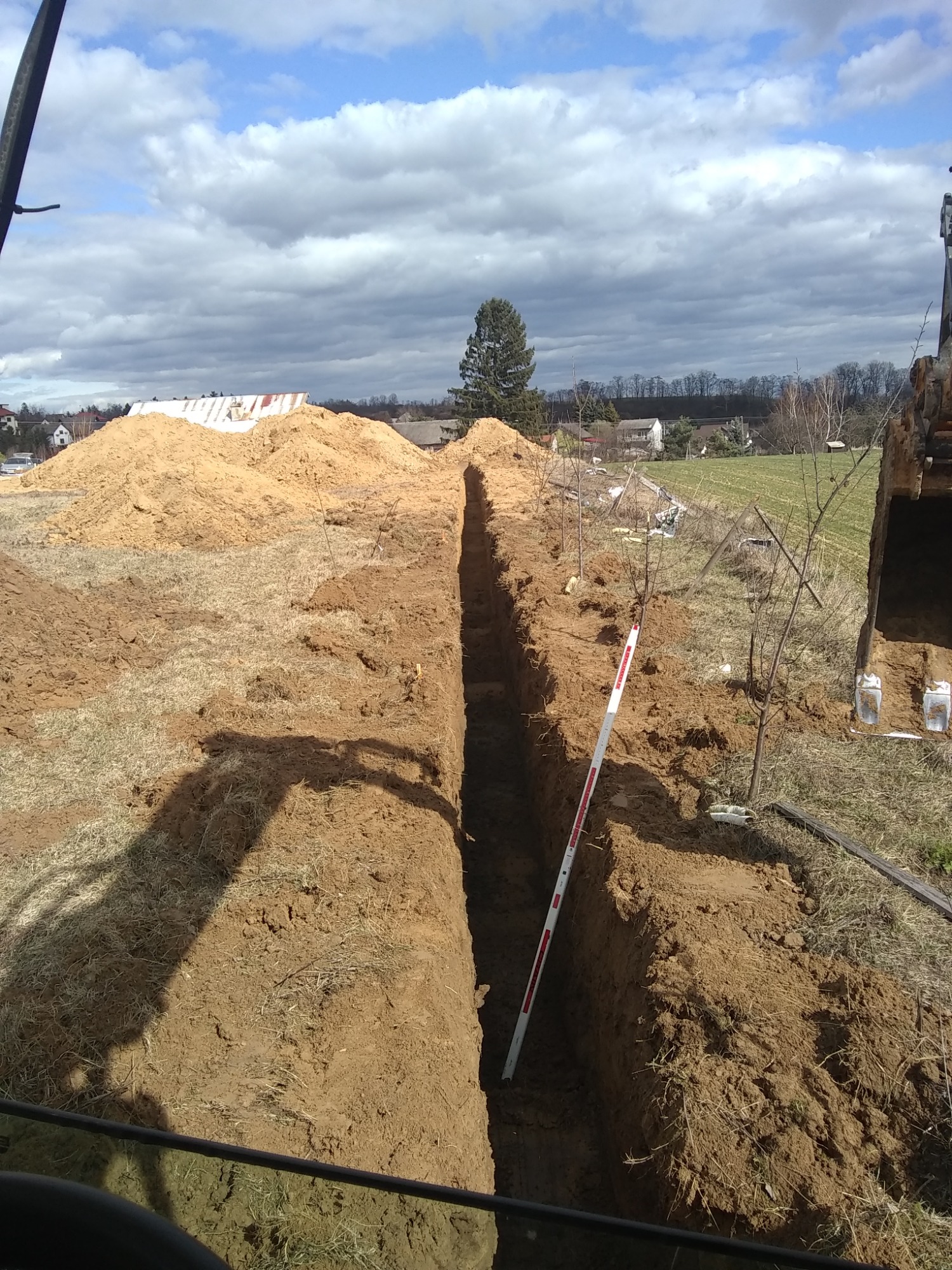 Wykopany wąski rów w ziemi z wystającą miarką pionową, w tle hałda ziemi i zabudowania wiejskie pod pochmurnym niebem.