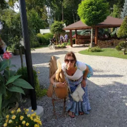 Uśmiechnięta kobieta w okularach przeciwsłonecznych pozuje obejmując metalową rzeźbę ptaka w ogrodzie z altaną i innymi osobami w tle.