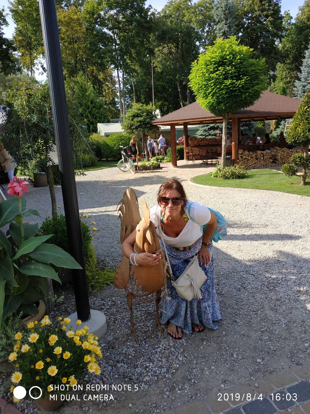 Uśmiechnięta kobieta w okularach przeciwsłonecznych pozuje obejmując metalową rzeźbę ptaka w ogrodzie z altaną i innymi osobami w tle.