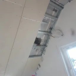 Poddasze w trakcie ocieplania, widoczna wełna mineralna, folia paroizolacyjna, profile metalowe i płyty gipsowo-kartonowe na skośnej ścianie, okno w tle.