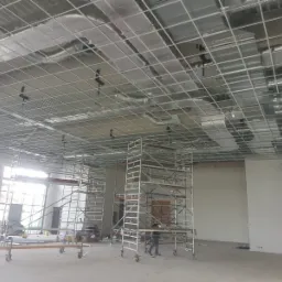 Montaż metalowej konstrukcji sufitu podwieszanego z widocznymi instalacjami wentylacyjnymi i ociepleniem, wsparty na rusztowaniach w przestronnym, jasnym pomieszczeniu w trakcie prac wykończeniowych.