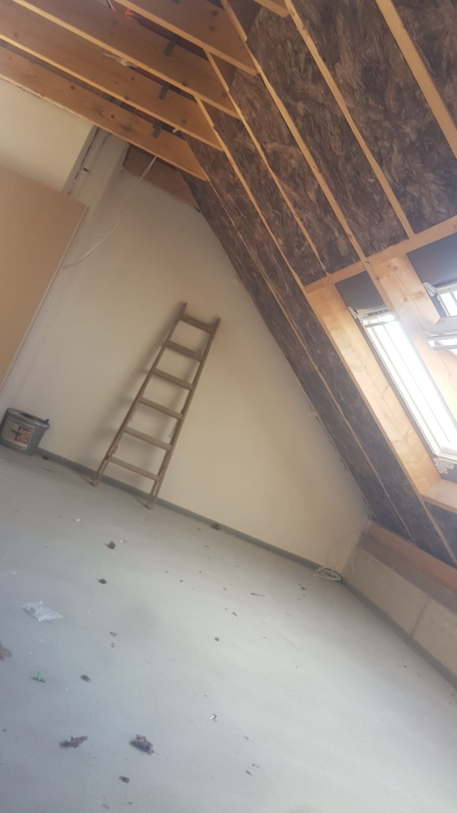 Poddasze w trakcie ocieplania wełną mineralną, widoczna drewniana konstrukcja dachu, drabina oparta o ścianę, okno dachowe i resztki materiałów na podłodze.