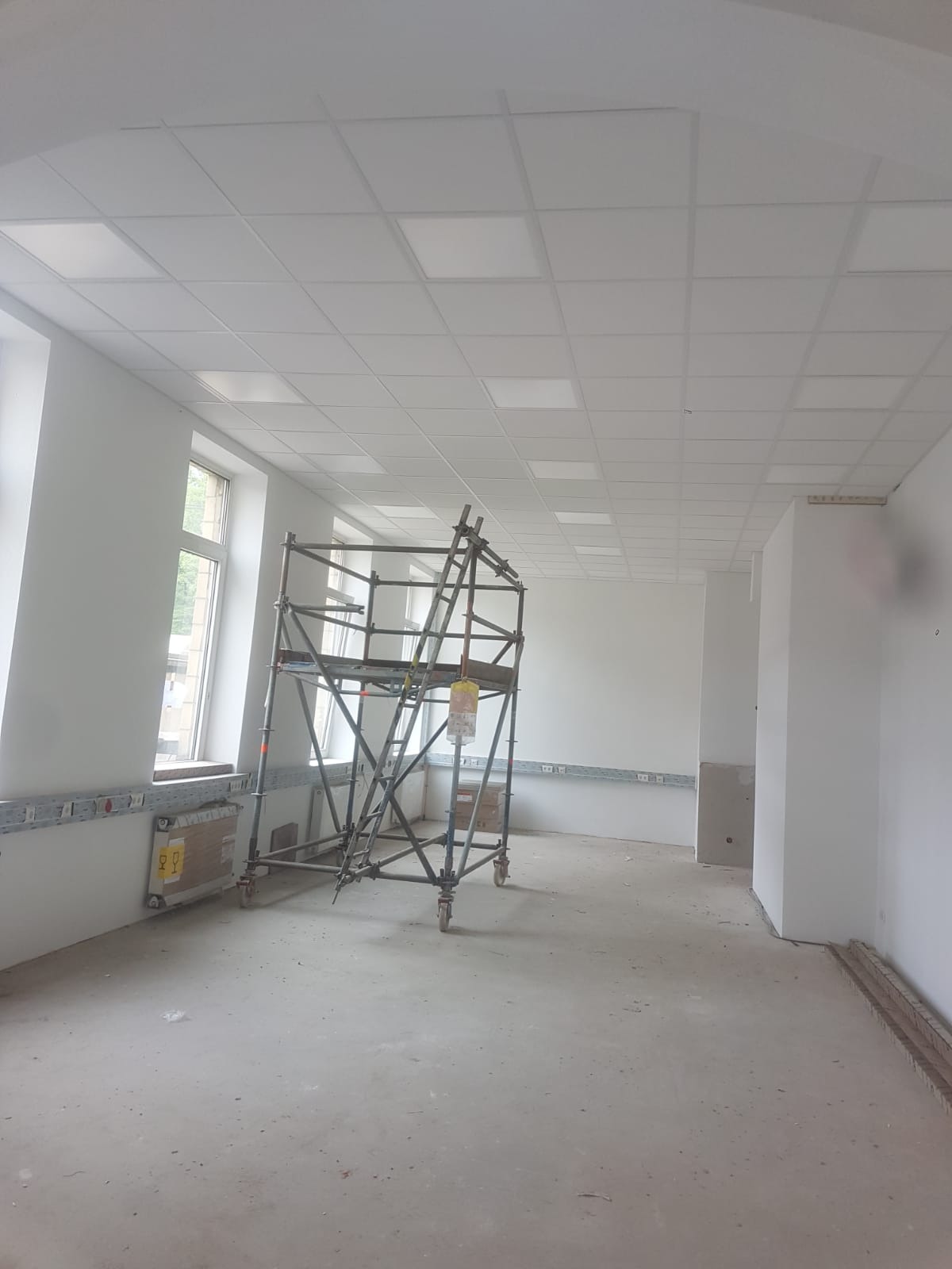 Wnętrze w trakcie prac wykończeniowych: widoczne rusztowanie na kółkach, świeżo pomalowane ściany na biało, sufit kasetonowy z wbudowanym oświetleniem oraz niezabezpieczona podłoga.