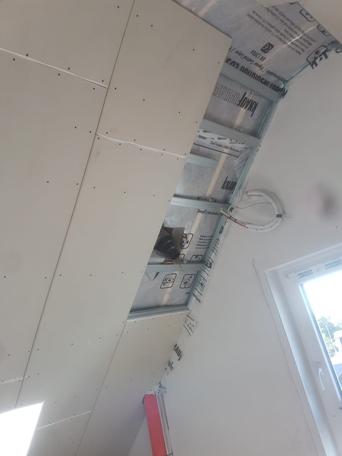 Poddasze w trakcie ocieplania, widoczna wełna mineralna, folia paroizolacyjna, profile metalowe i płyty gipsowo-kartonowe na skośnej ścianie, okno w tle.