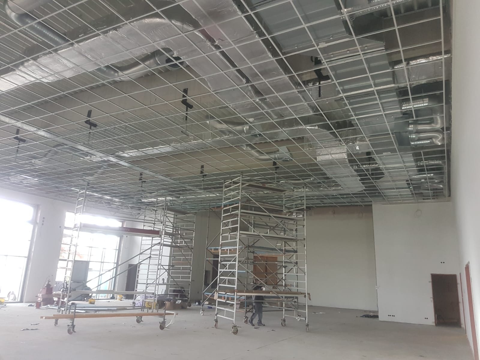 Montaż metalowej konstrukcji sufitu podwieszanego z widocznymi instalacjami wentylacyjnymi i ociepleniem, wsparty na rusztowaniach w przestronnym, jasnym pomieszczeniu w trakcie prac wykończeniowych.