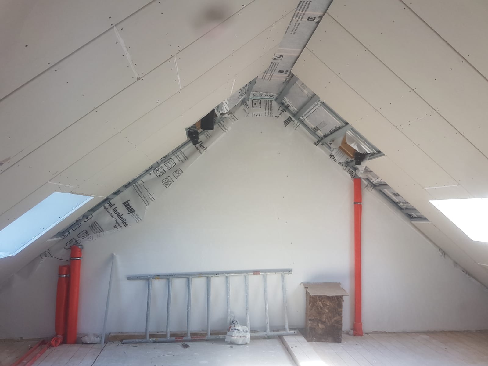 Poddasze w trakcie prac wykończeniowych, widoczne ocieplenie wełną mineralną zabezpieczone folią paroizolacyjną Knauf, płyty gipsowo-kartonowe na skosach, fragment okna dachowego, pomarańczowe rury...