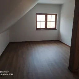 Poddasze z drewnianą podłogą, widoczne drewniane okno w białej ścianie oraz drewniany słup konstrukcyjny. Ściany pomalowane na biało.