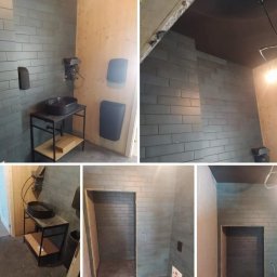 Ad-Serwis - Wnętrze łazienki w trakcie prac wykończeniowych: szare, podłużne płytki na ścianach, drewniane panele, czarna umywalka na metalowym stelażu z półką, widoczne elementy instalacji elektrycznej...