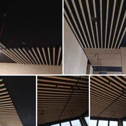 Ad-Serwis - Drewniany ażurowy sufit z listew na czarnym tle, widoczne przewody instalacji elektrycznej, surowe wykończenie ścian z płyt drewnianych.