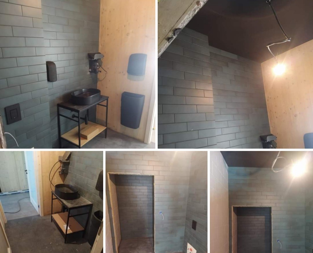 Wnętrze łazienki w trakcie prac wykończeniowych: szare, podłużne płytki na ścianach, drewniane panele, czarna umywalka na metalowym stelażu z półką, widoczne elementy instalacji elektrycznej...