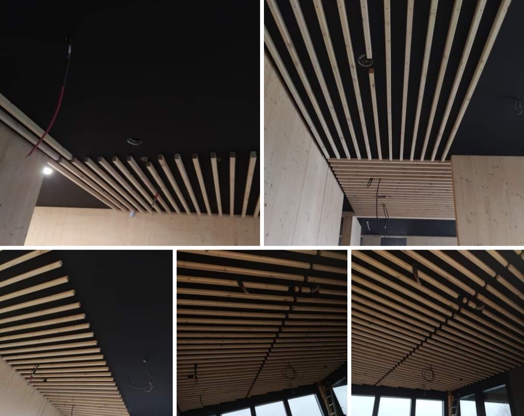 Drewniany ażurowy sufit z listew na czarnym tle, widoczne przewody instalacji elektrycznej, surowe wykończenie ścian z płyt drewnianych.