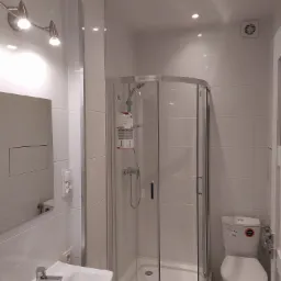 Jasna łazienka po remoncie z kabiną prysznicową narożną, umywalką z szafką i toaletą, wykończona białymi płytkami, oświetlona punktowymi lampami sufitowymi i kinkietem nad lustrem.