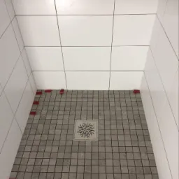 Płytki podłogowe w kabinie prysznicowej, szare kwadratowe mozaiki z odpływem liniowym w kształcie rozety, białe płytki ścienne z czerwonymi krzyżykami dystansowymi przy podłodze.