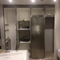 Wnętrze kuchni w trakcie montażu zabudowy: widoczne szafki z szarymi ramami, półki z akcesoriami i sprzętem, oraz lodówka w metalowej obudowie. Podłoga częściowo zabezpieczona. Na pierwszym planie...