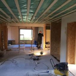 Leon - Wnętrze domu w trakcie prac wykończeniowych: widoczne drewniane elementy konstrukcyjne ścian i stropu, podłoga z przewodami, kompresor, drabina i sylwetka osoby w tle.