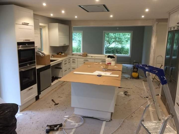 Wnętrze kuchni w trakcie remontu: białe szafki, wyspa kuchenna zabezpieczona kartonem, widoczne narzędzia, drabina oparta o lodówkę, gruz na podłodze, okno z widokiem na zieleń.