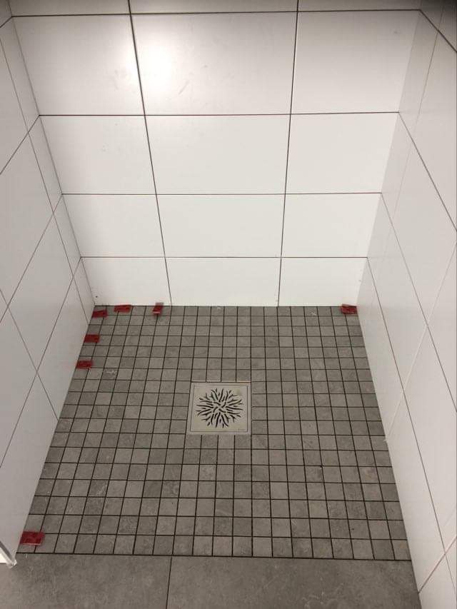 Płytki podłogowe w kabinie prysznicowej, szare kwadratowe mozaiki z odpływem liniowym w kształcie rozety, białe płytki ścienne z czerwonymi krzyżykami dystansowymi przy podłodze.