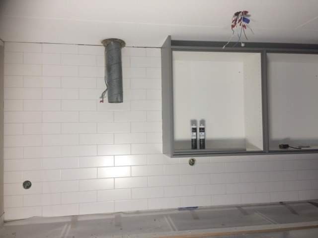 Ściana wyłożona białymi, prostokątnymi płytkami, z widocznym przewodem wentylacyjnym i częściowo zamontowanymi szafkami kuchennymi w szarych ramach, dwie puszki z produktami na półce, widoczne...