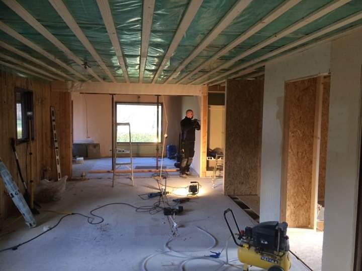 Wnętrze w trakcie remontu: widoczne drewniane legary pod sufitem z folią, ściany w surowym stanie, narzędzia i kompresor na podłodze, majster w tle.