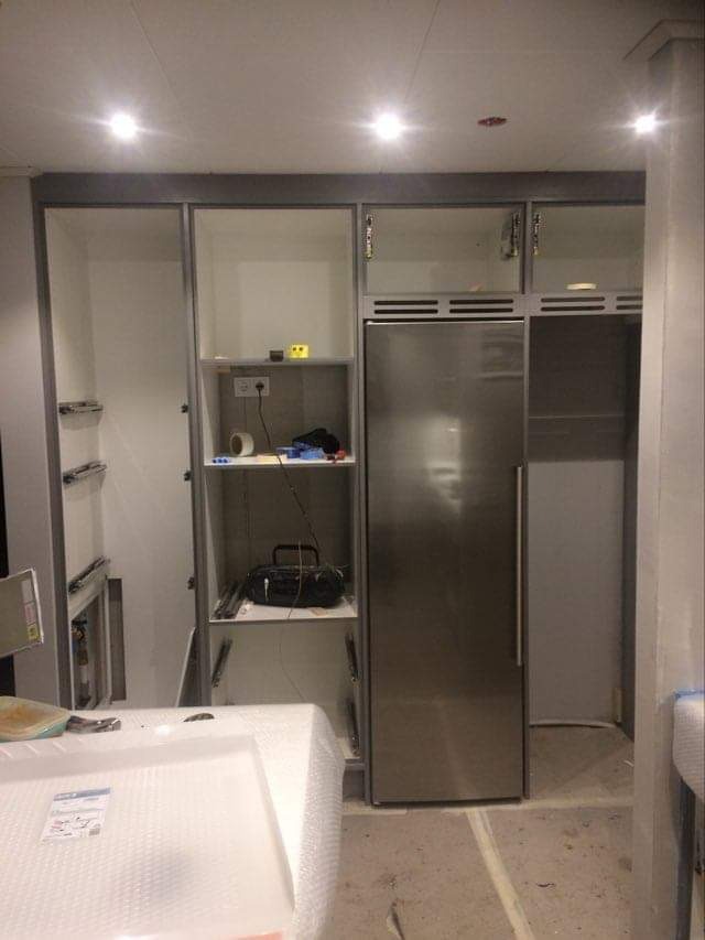 Wnętrze kuchni w trakcie montażu zabudowy: widoczne szafki z szarymi ramami, półki z akcesoriami i sprzętem, oraz lodówka w metalowej obudowie. Podłoga częściowo zabezpieczona. Na pierwszym planie...