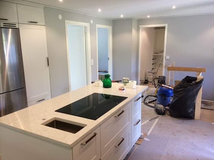 Kuchnia w trakcie prac wykończeniowych: wyspa kuchenna z płytą indukcyjną i zlewem, widoczne narzędzia, odkurzacz i ślady po malowaniu na podłodze.