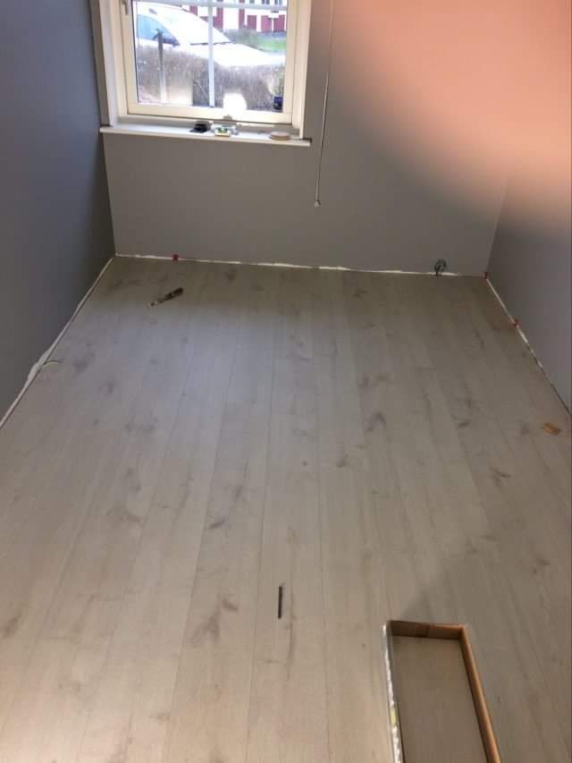 Jasne panele podłogowe ułożone w pokoju z szarymi ścianami, widoczne plastikowe kliny dystansowe przy ścianach i otwór na rurę.