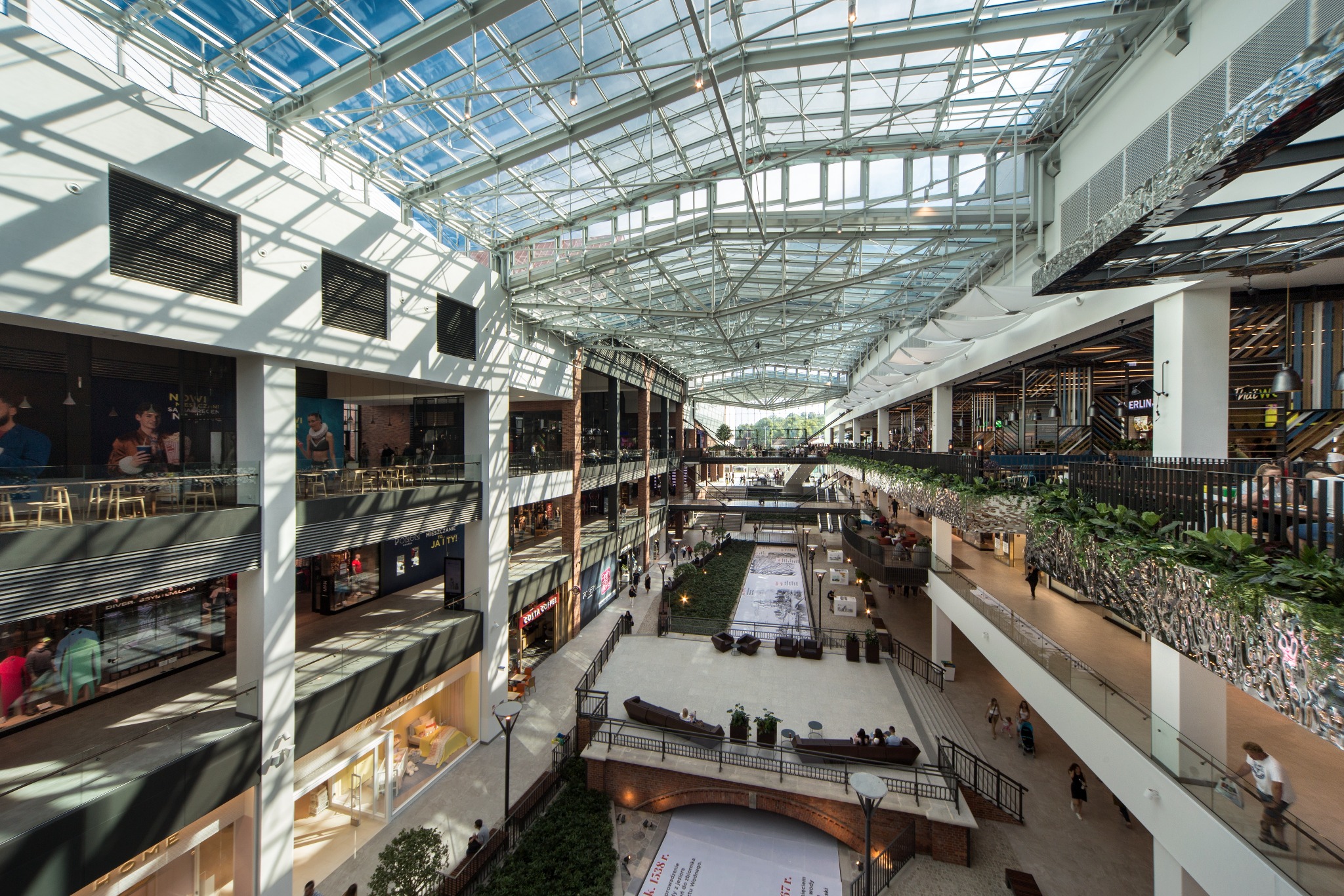 Wielopoziomowe wnętrze centrum handlowego z przeszklonym dachem, widok z góry na alejki, sklepy i strefy wypoczynku z roślinnością.