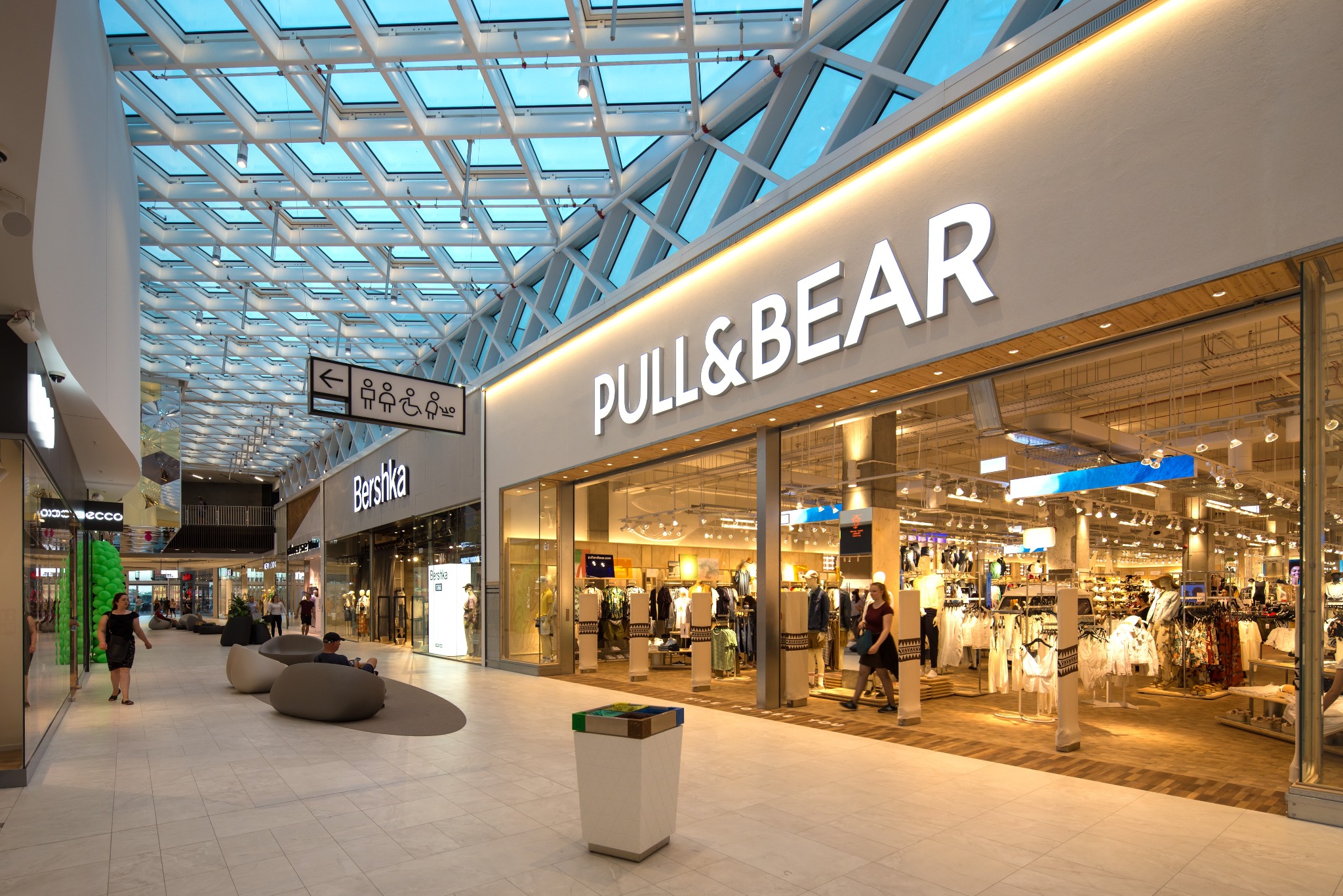 Wnętrze Forum Gdańsk z widokiem na sklepy Pull&Bear i Bershka pod przeszklonym dachem, z nowoczesnymi miejscami do siedzenia i koszem na śmieci na pierwszym planie.