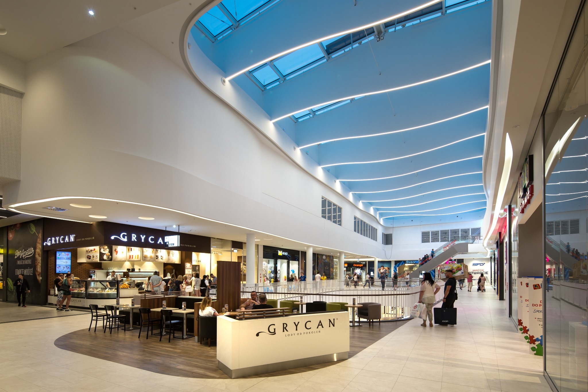 Wnętrze nowoczesnego centrum handlowego Forum Gdańsk z widoczną kawiarnią Grycan, oświetlonym sufitem z niebieskimi świetlikami i białymi liniami LED oraz licznymi sklepami w tle.