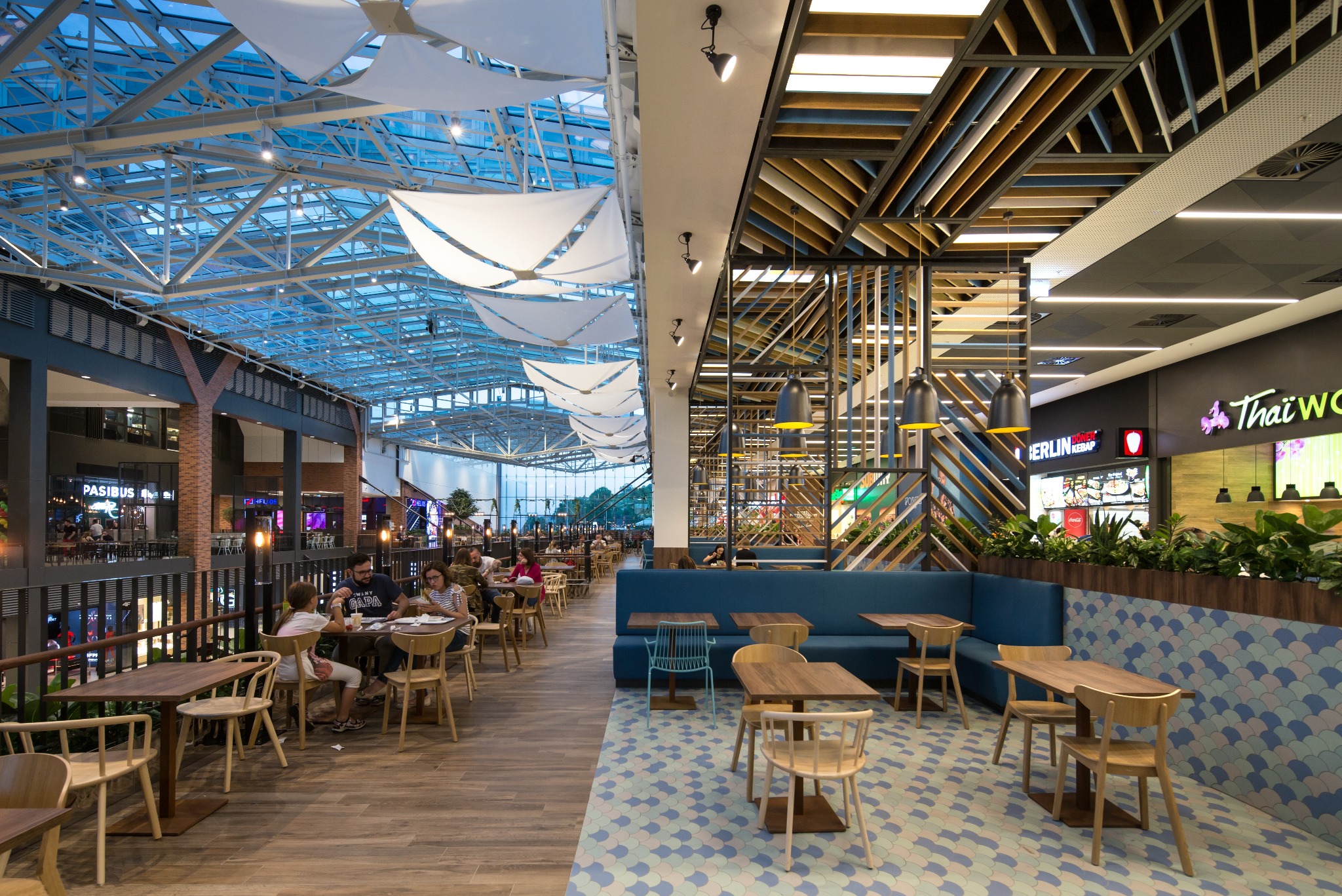 Wnętrze centrum handlowego Forum Gdańsk: drewniane podłogi, szklany sufit z białymi żaglami, strefa restauracyjna z różnorodnymi miejscami do siedzenia i dekoracyjnymi elementami architektonicznymi.