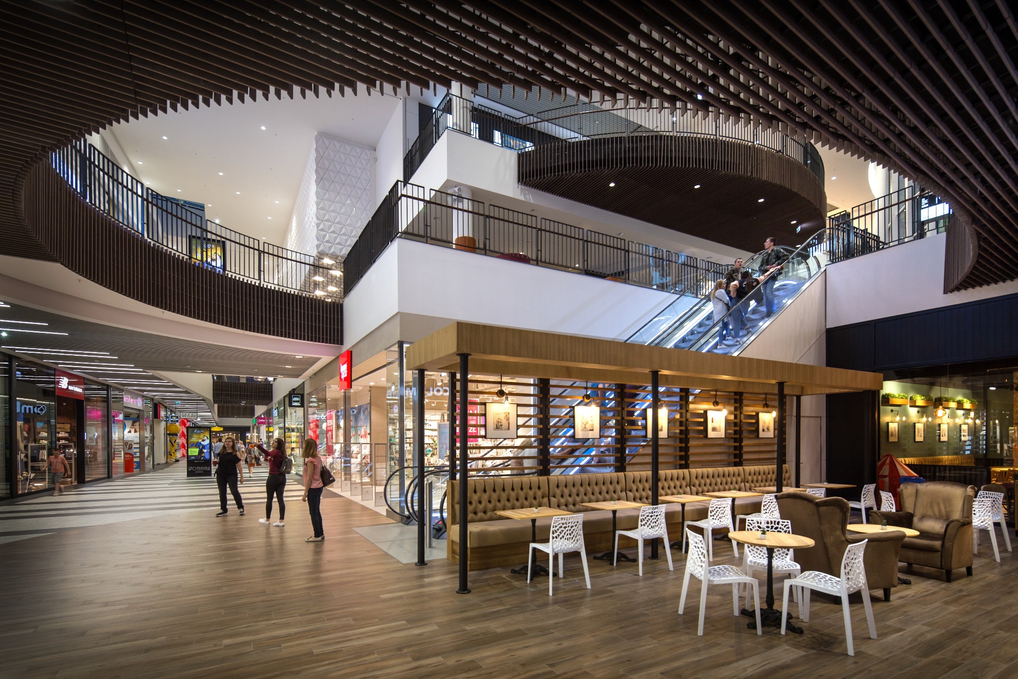 Wnętrze centrum handlowego Forum Gdańsk: widok na kawiarnię z beżową tapicerowaną ławą i białymi krzesłami, schody ruchome w tle, drewniana podłoga i ażurowy sufit.