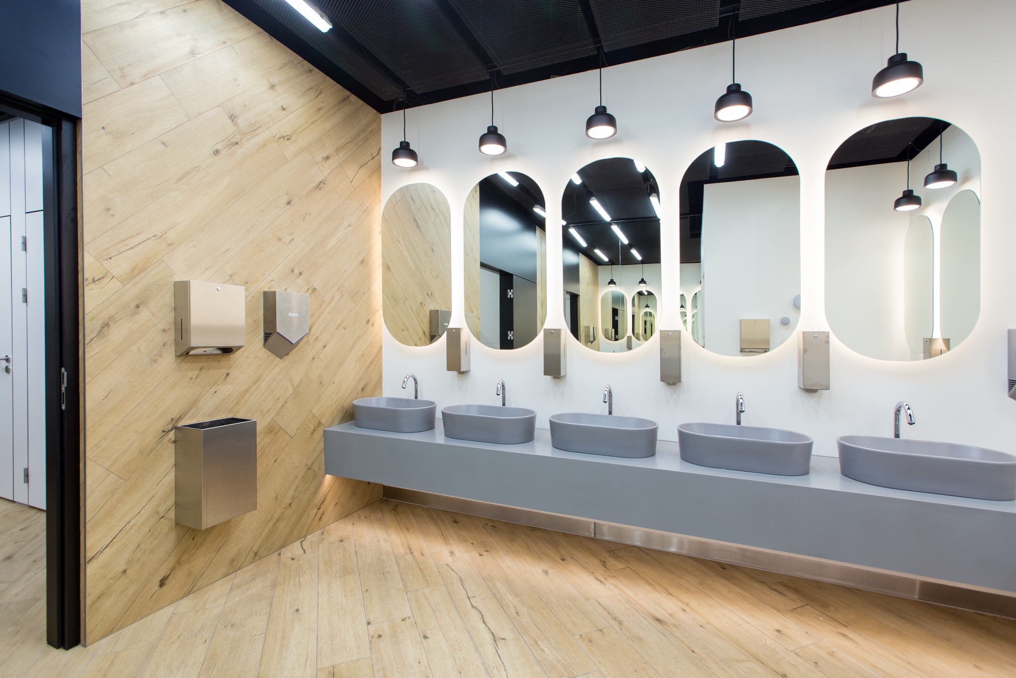 Wnętrze nowoczesnej toalety publicznej z drewnianą okładziną ścienną, podświetlanymi owalnymi lustrami, szarymi umywalkami i metalowymi dozownikami.