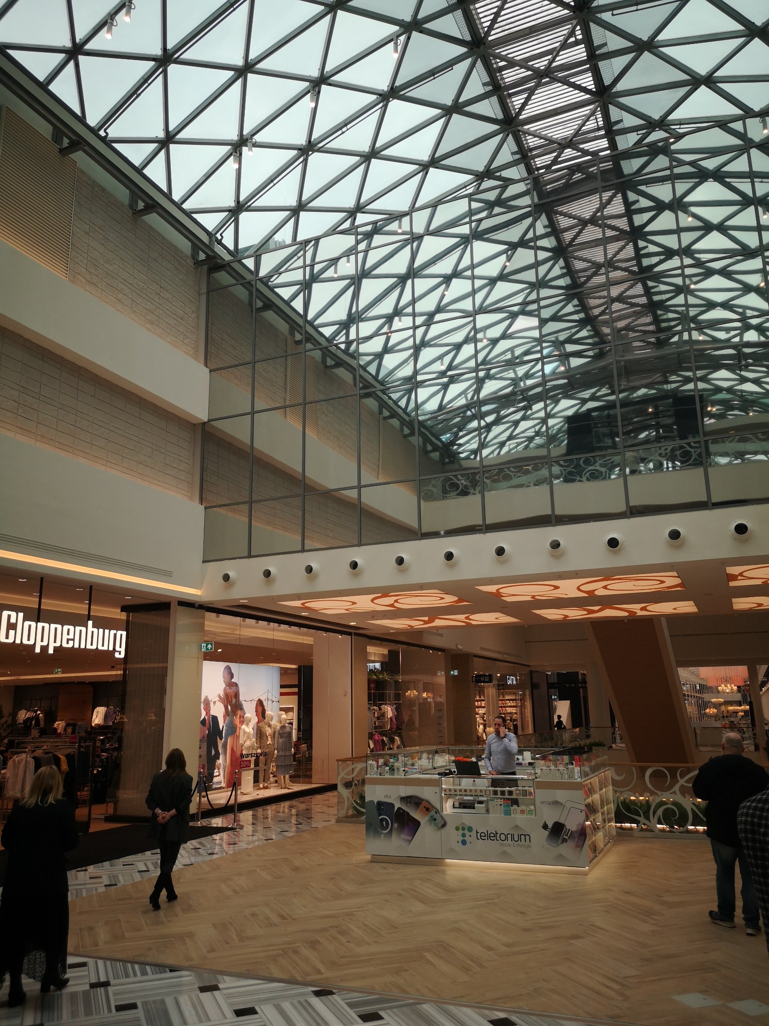 Wnętrze centrum handlowego z przeszklonym, trójkątnym dachem, widok na sklepy Cloppenburg i Teletorium, podłoga wyłożona parkietem w jodełkę.
