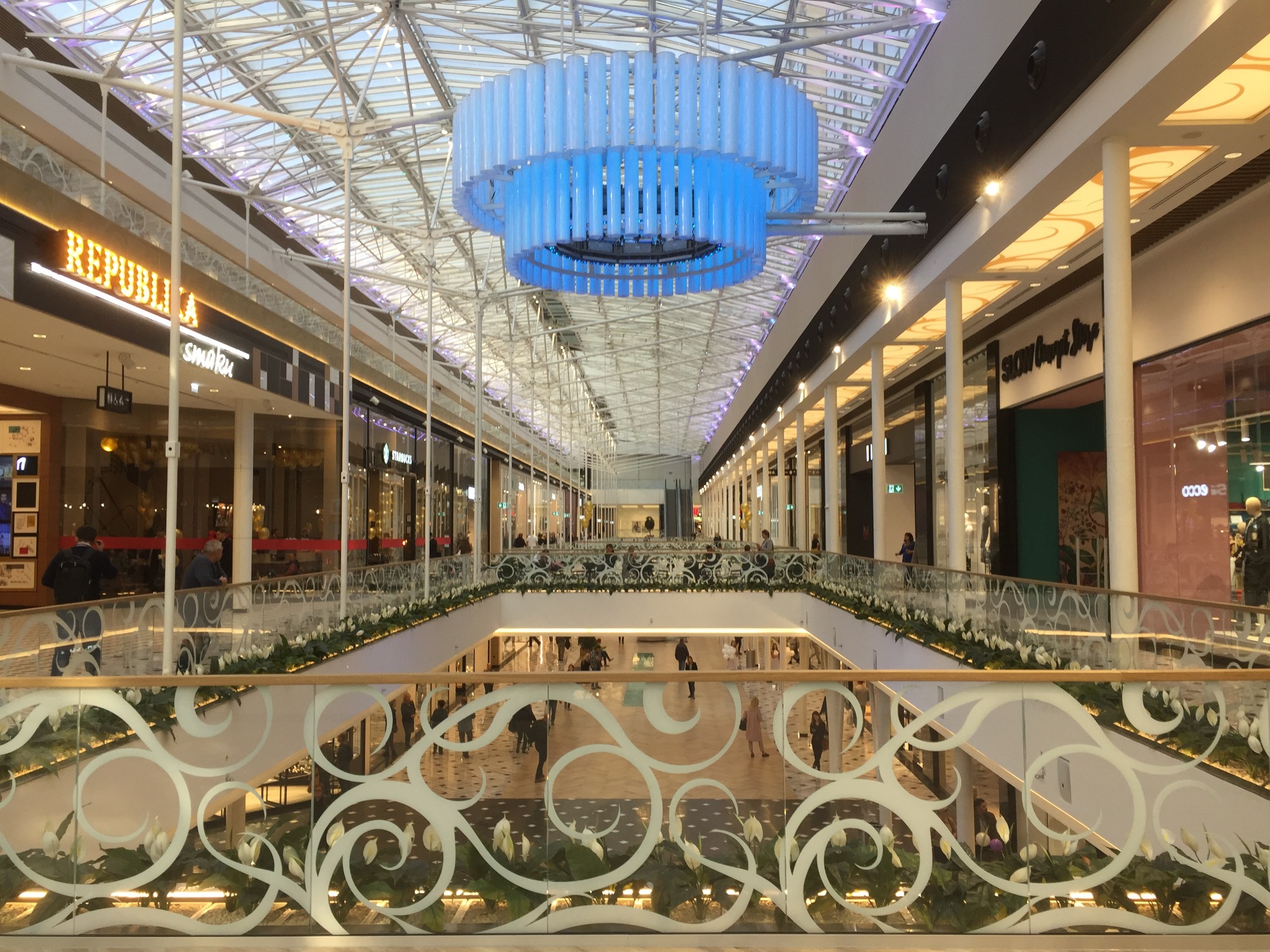 Wnętrze galerii handlowej z dwupoziomowym układem, szklanymi balustradami z dekoracyjnymi wzorami roślinnymi, widoczne sklepy i duża, okrągła lampa sufitowa w odcieniach błękitu.