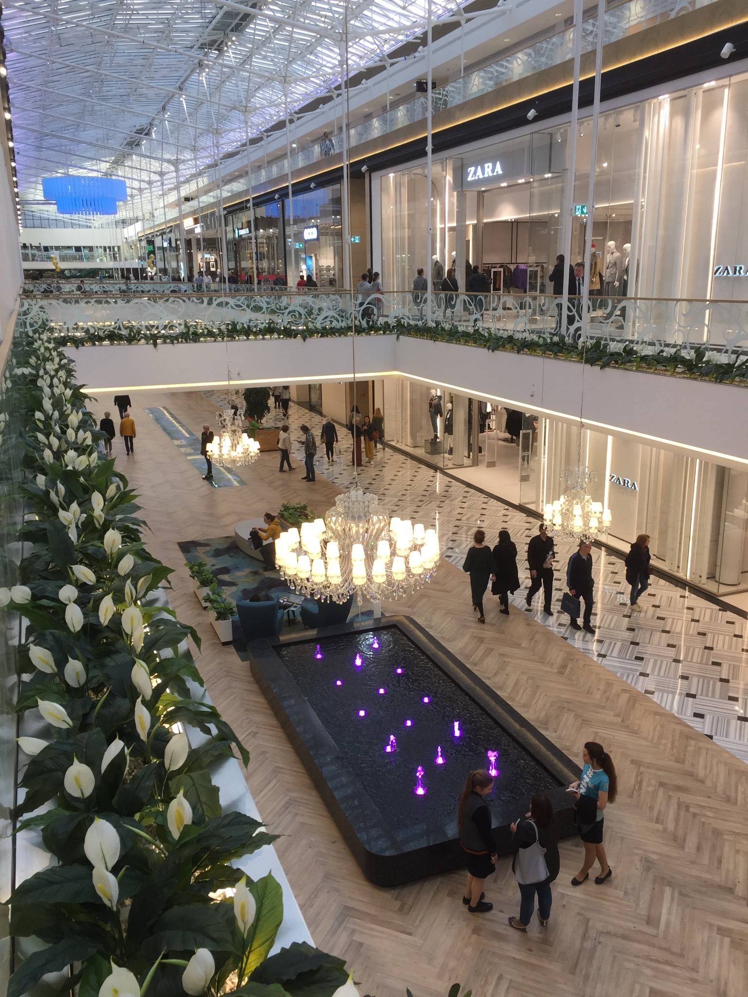 Wnętrze centrum handlowego z perspektywy antresoli, widoczne sklepy Zara, dekoracyjne rośliny, fontanna z podświetleniem LED i przechodzący ludzie.