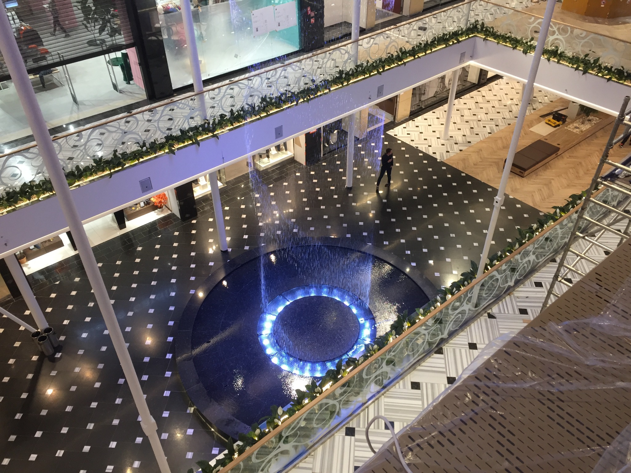 Wnętrze galerii handlowej z fontanną w kształcie okręgu podświetloną na niebiesko, widok z góry, balustrady z roślinami, osoba spacerująca po czarnej posadzce w białe kwadraty.