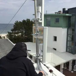Instalator w czarnej bluzie i czapce instaluje antenę na dachu budynku z widokiem na morze i plażę. Antena przymocowana do metalowej konstrukcji, widoczne kable i elementy montażowe.