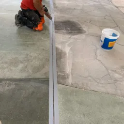 Pracownik na kolanach aplikuje uszczelniacz do szczeliny dylatacyjnej w szarej posadzce przemysłowej, widoczny biały pojemnik z niebieskim logo i ciemne plamy na betonie.