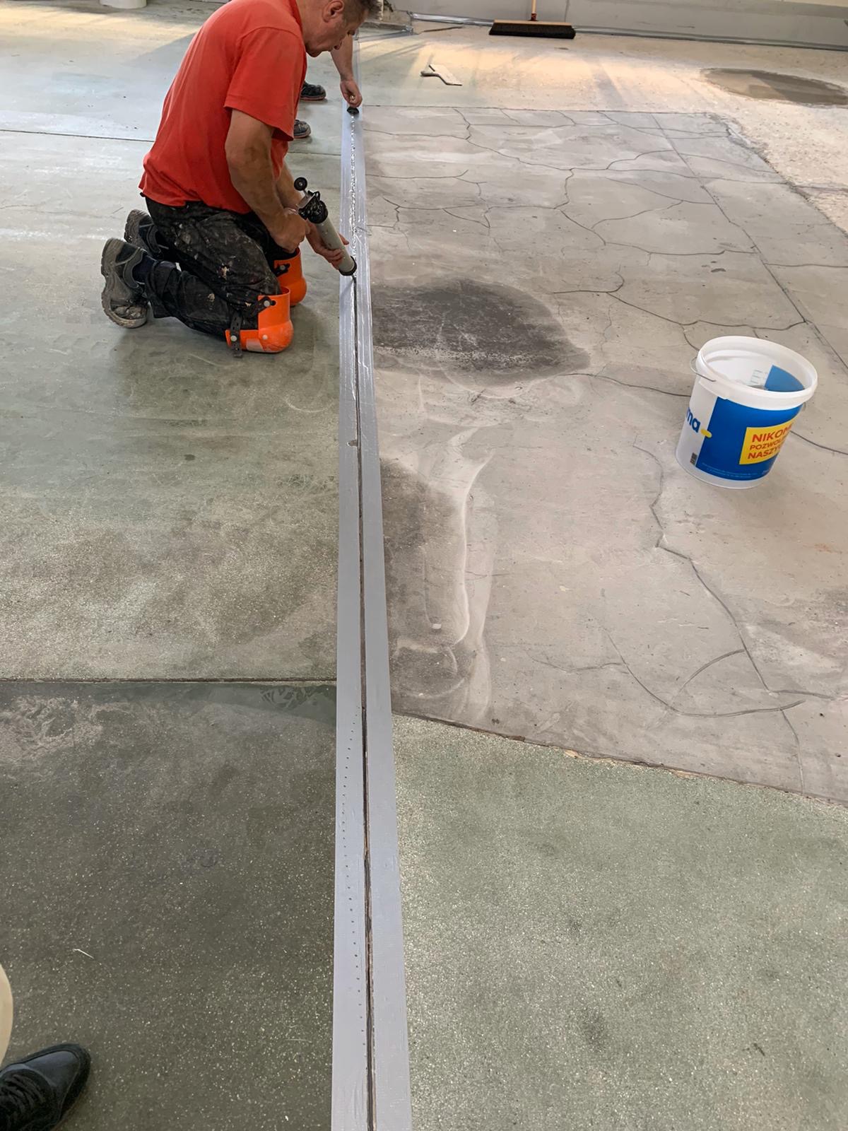 Pracownik na kolanach aplikuje uszczelniacz do szczeliny dylatacyjnej w szarej posadzce przemysłowej, widoczny biały pojemnik z niebieskim logo i ciemne plamy na betonie.