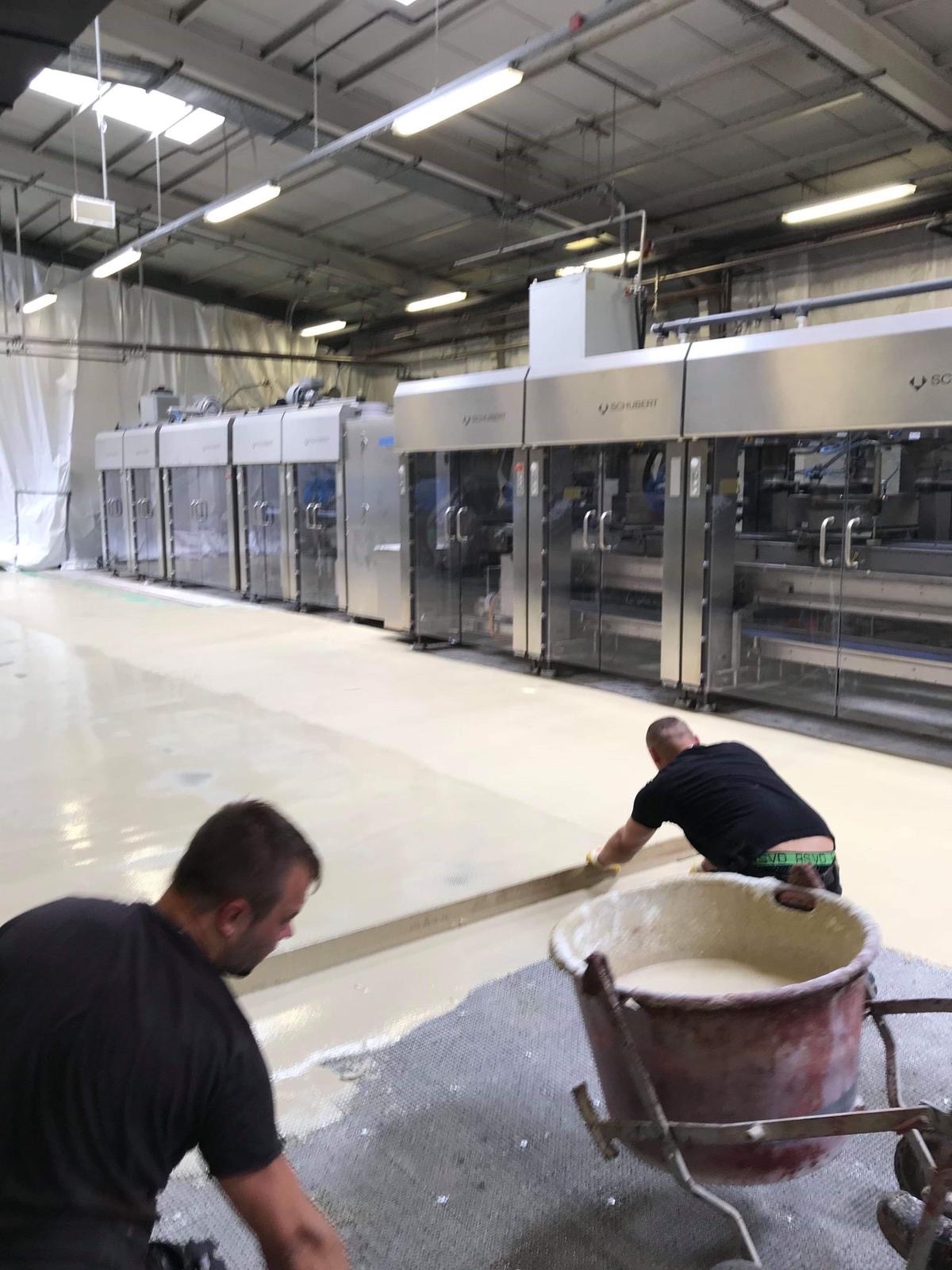 Pracownicy w trakcie wylewania jasnej, samopoziomującej posadzki w hali produkcyjnej z widocznymi maszynami Schubert w tle, używając metalowej listwy i wiadra z zaprawą.