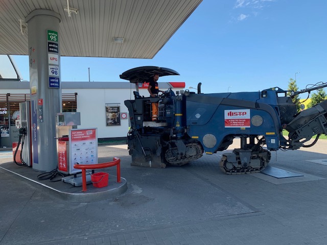 Frezarka do asfaltu na kostce brukowej stacji benzynowej, operator w kabinie, widoczny znak firmowy ISTP, czerwone wiadro obok dystrybutora paliwa.