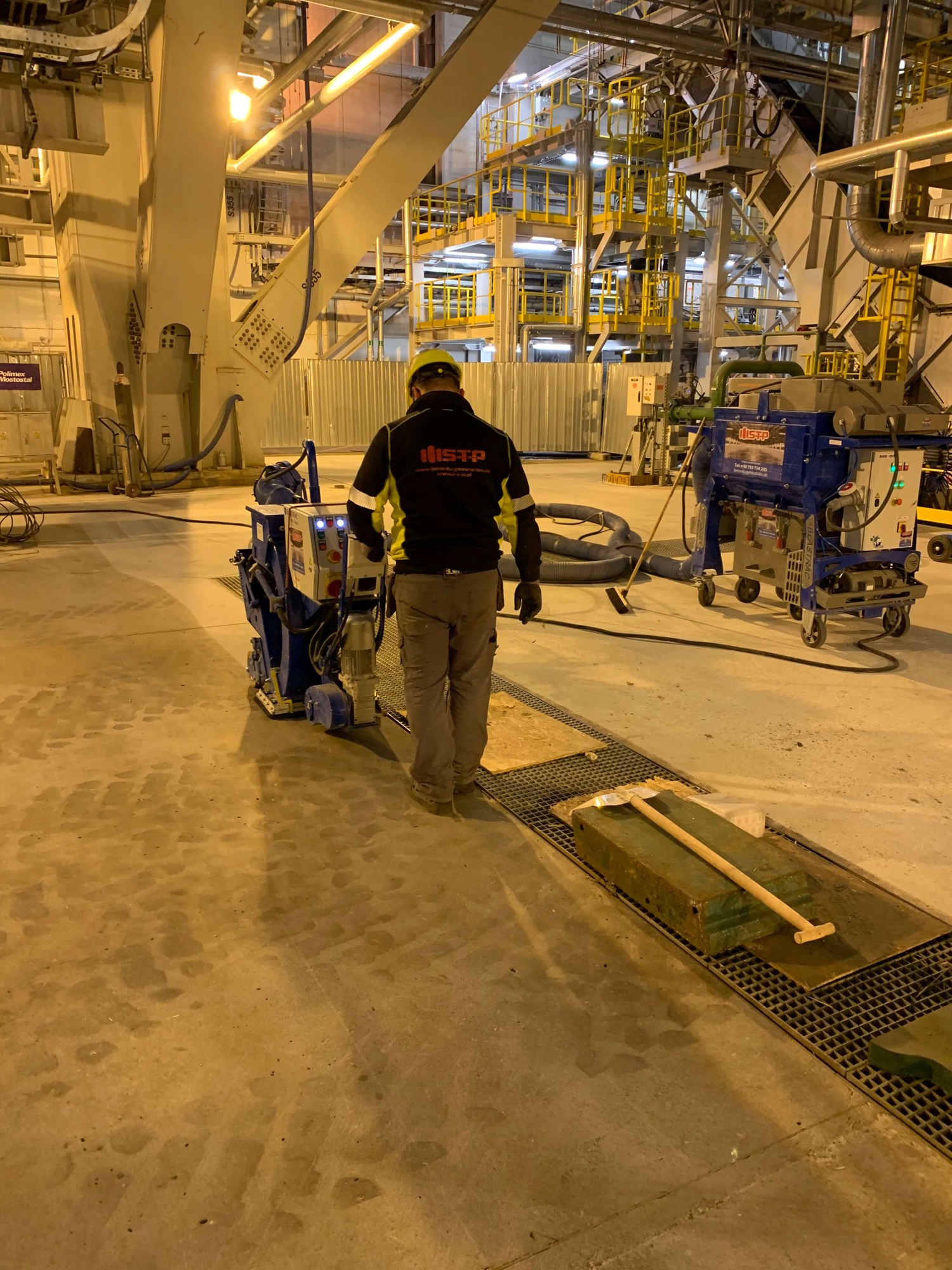 Pracownik w kasku obsługuje maszynę do czyszczenia posadzek przemysłowych Blastrac w hali fabrycznej, widoczne ślady po czyszczeniu oraz kratka ściekowa z leżącym narzędziem.
