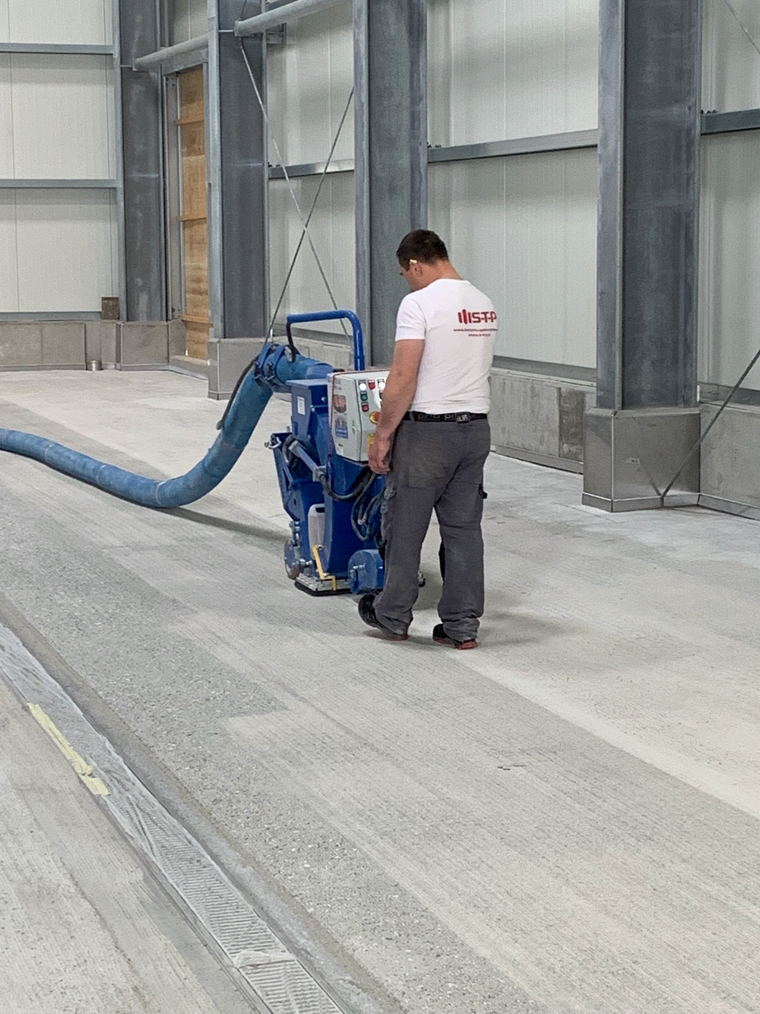 Pracownik w słuchawkach obsługuje maszynę do obróbki betonu na posadzce w hali przemysłowej, widoczny wąż odkurzacza i stalowa konstrukcja budynku.