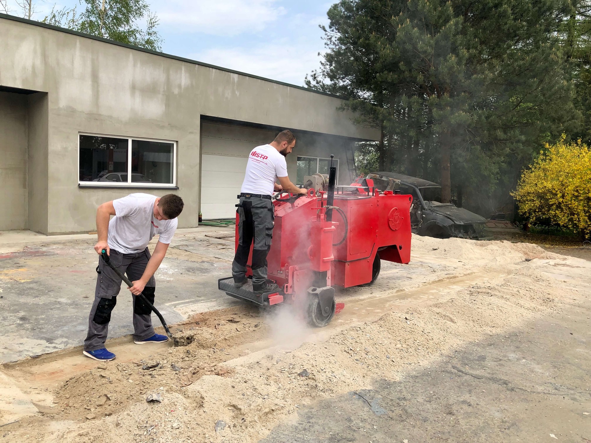 Dwóch pracowników wykonuje nacięcia w betonowej posadzce za pomocą czerwonej maszyny frezującej, unoszący się pył, budynek w tle.