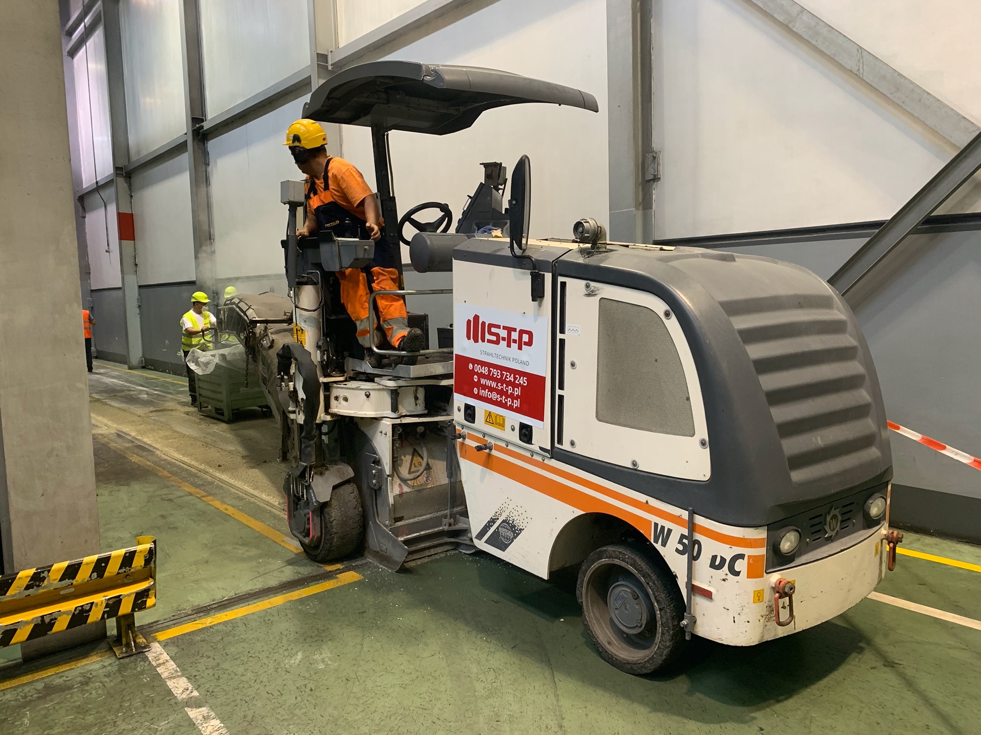 Operator w żółtym kasku obsługuje frezarkę do posadzek w hali przemysłowej, widoczny napis 'ISTP Strahltechnik Poland' na maszynie, w tle pracownik w kamizelce odblaskowej.