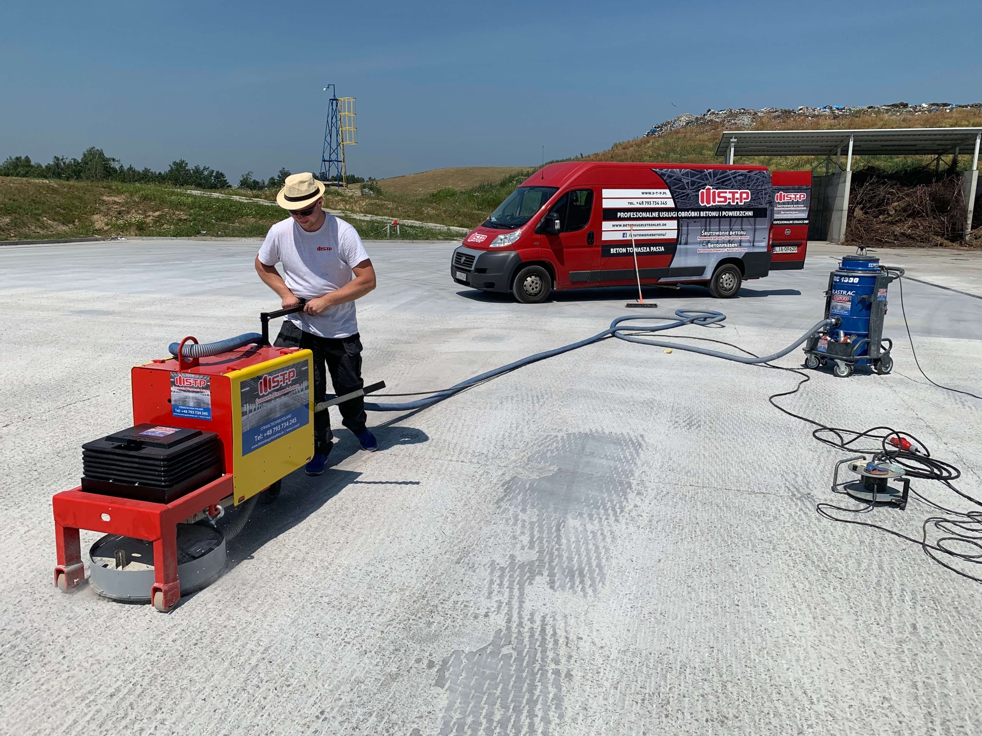 Pracownik w kapeluszu obsługuje szlifierkę do betonu na dużej betonowej powierzchni, w tle czerwony van z reklamą firmy i wysypisko śmieci.